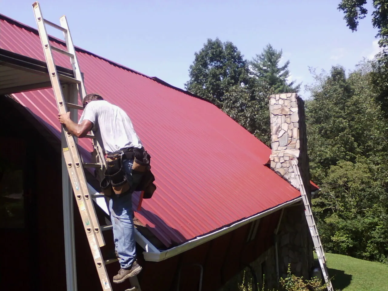Roofer on a ladder beside a red metal roof and stone chimney — Soffit & Fascia Repair by Roofing Brothers in Brinkhaven